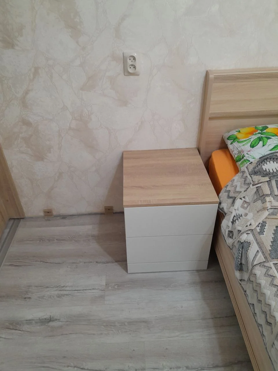 Прикроватная тумба рядом с деревянным изголовьем и краем спального места